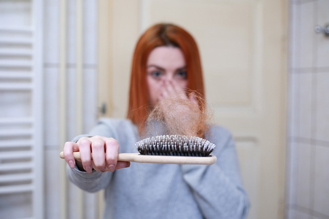 Eine Person mit roten Haaren hält eine Haarbürste, in der deutlich mehr Haarsträhnen stecken, und bedeckt mit der anderen Hand ihren Mund, was möglicherweise ihre Besorgnis über Haarausfall zum Ausdruck bringt, und steht in einem Badezimmer.